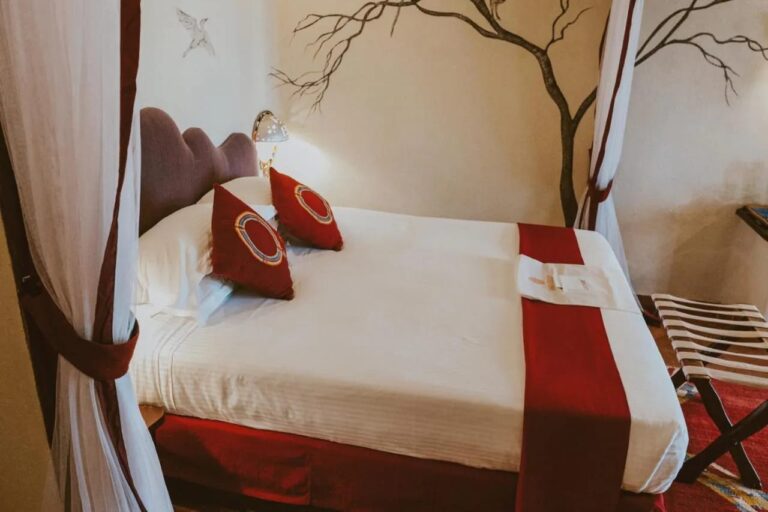 Duże łóżko z czerwonymi poduszkami między dwiema białymi zasłonami, przed drzewem namalowanym na ścianie