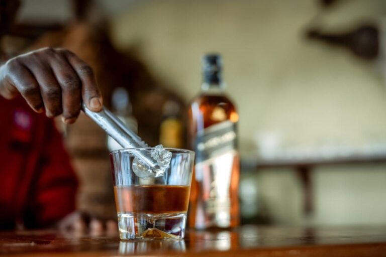 Dłoń wkłada kostkę lodu do szklanki whisky na ladzie baru