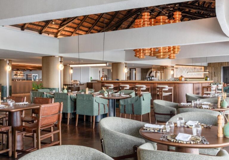 Restauracja z zielonymi fotelami przed długim barem, pod pomarańczowymi lampami wiszącymi