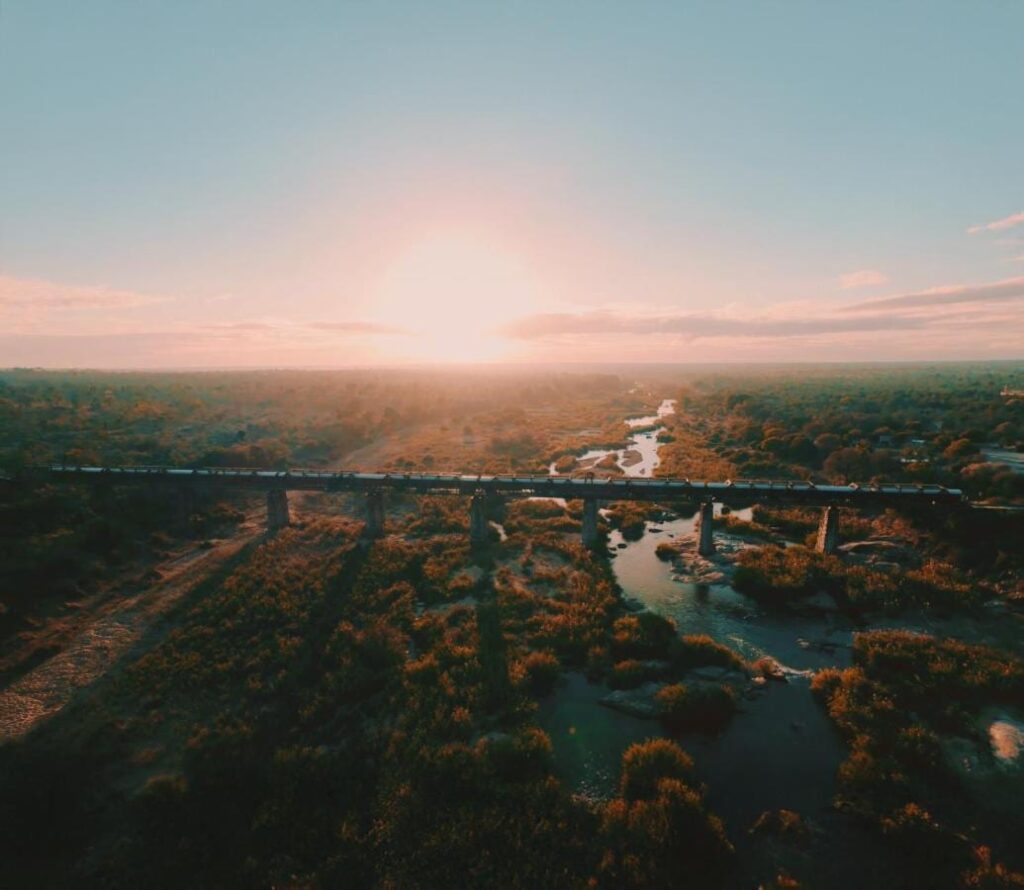 Pociąg Kruger Shalati na moście nad rzeką Selati z lotu ptaka, nad wodą i buszem, przy nisko wiszącym słońcu