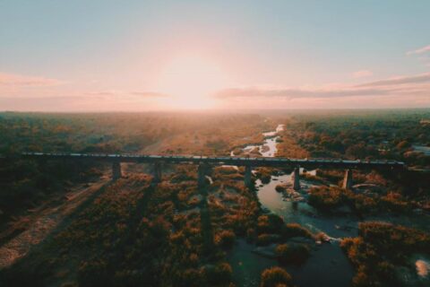 Pociąg Kruger Shalati na moście nad rzeką Selati z lotu ptaka, nad wodą i buszem, przy nisko wiszącym słońcu