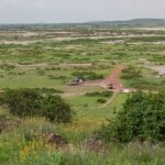 Widok z Observation Hill na zieloną równinę w plamach soli, czerwona droga gruntowa, trzy samochody 4x4 przy małym ochrowym budynku, w oddali niskie wzgórza