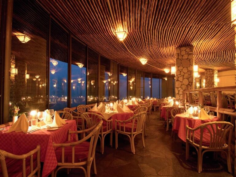 Sala restauracyjna oświetlona świecami, z czerwonymi obrusami i rattanowymi fotelami pod drewnianym sufitem