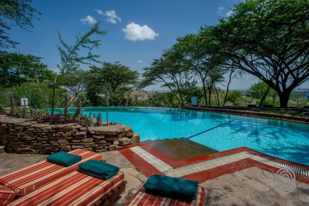 Basen bez krawędzi Serengeti Serena Safari Lodge z leżakami w czerwone paski i zielonymi ręcznikami, pod akacjami