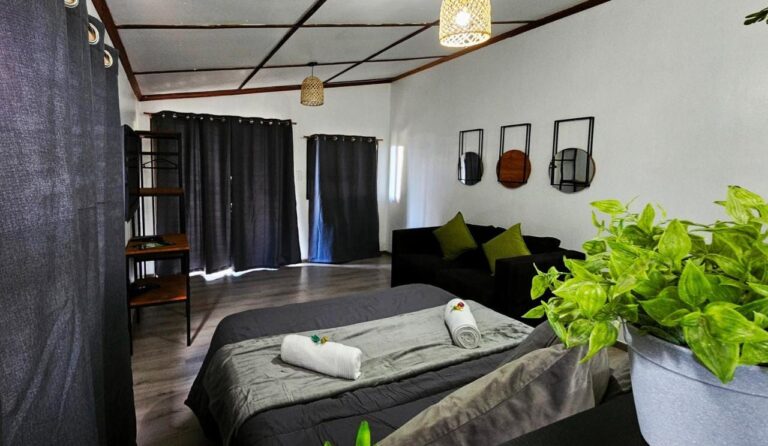 Duże szare łóżko ze zwiniętymi ręcznikami, czarna sofa i ciemne zasłony w studiu z sufitem w drewnianą kratę